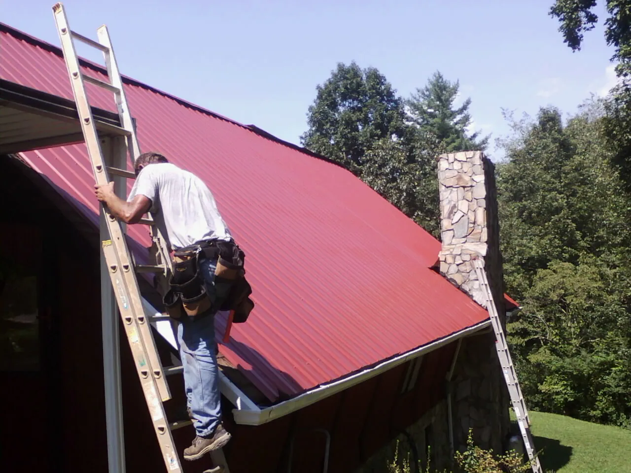 Roofer on a ladder beside a red metal roof and stone chimney — Soffit & Fascia Repair by Roofing Brothers in Topsham